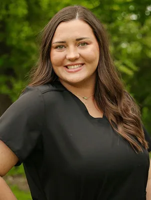 A smiling woman with long brown hair in black scrubs.